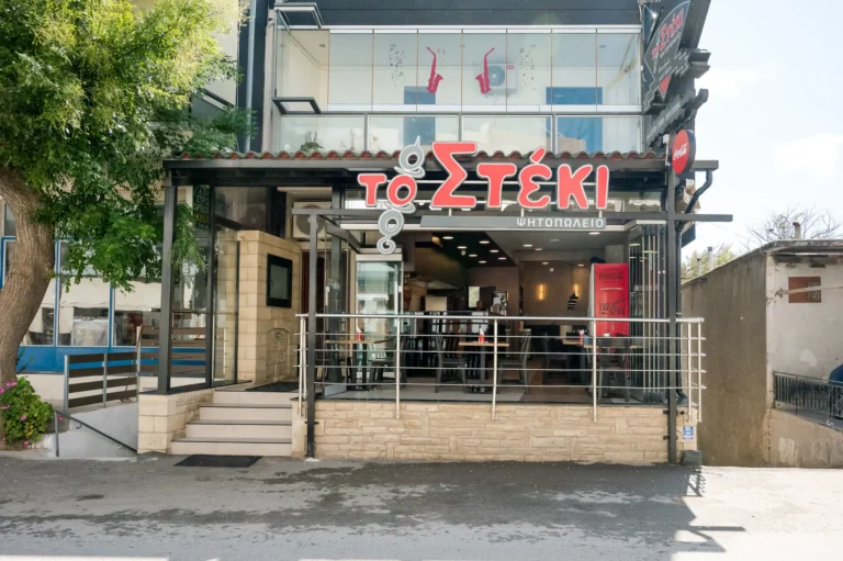 Η πρόσοψη του ψητοπωλείου Το Στέκι με την κόκκινη επιγραφή και τα τραπέζια στη βεράντα